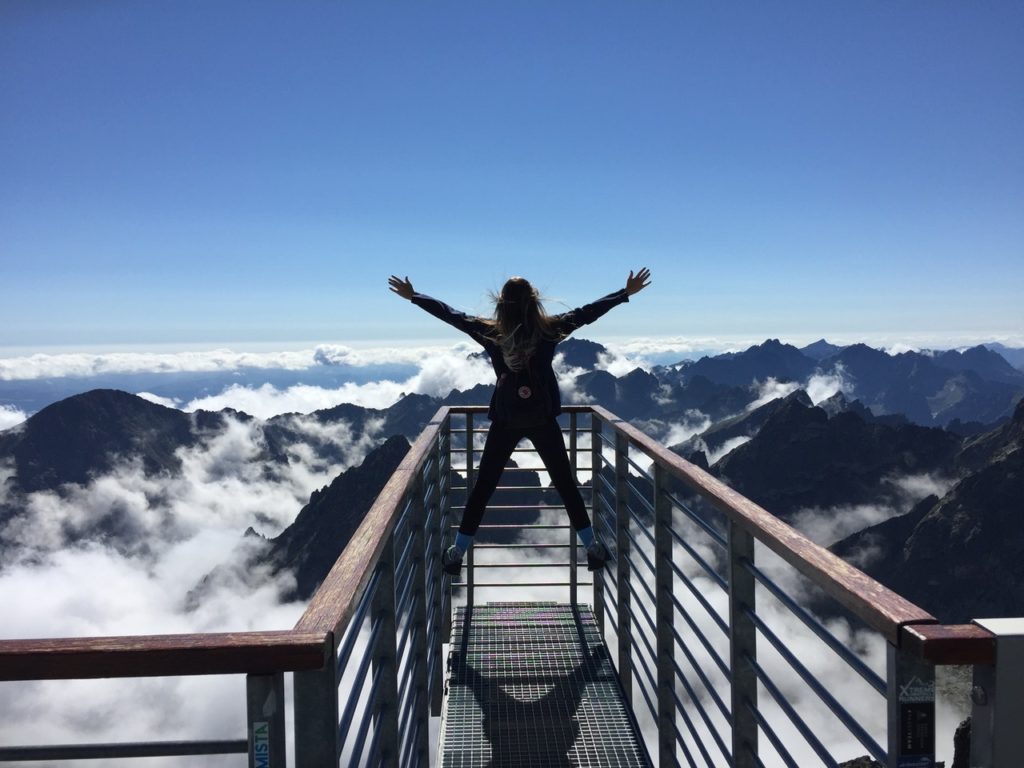 A woman with her arms stretched out to both sides has climbed up a guard rail of a platform above the clouds. In the far off distance you can see mountains peeking out from above the clouds all the way to the horizon. 